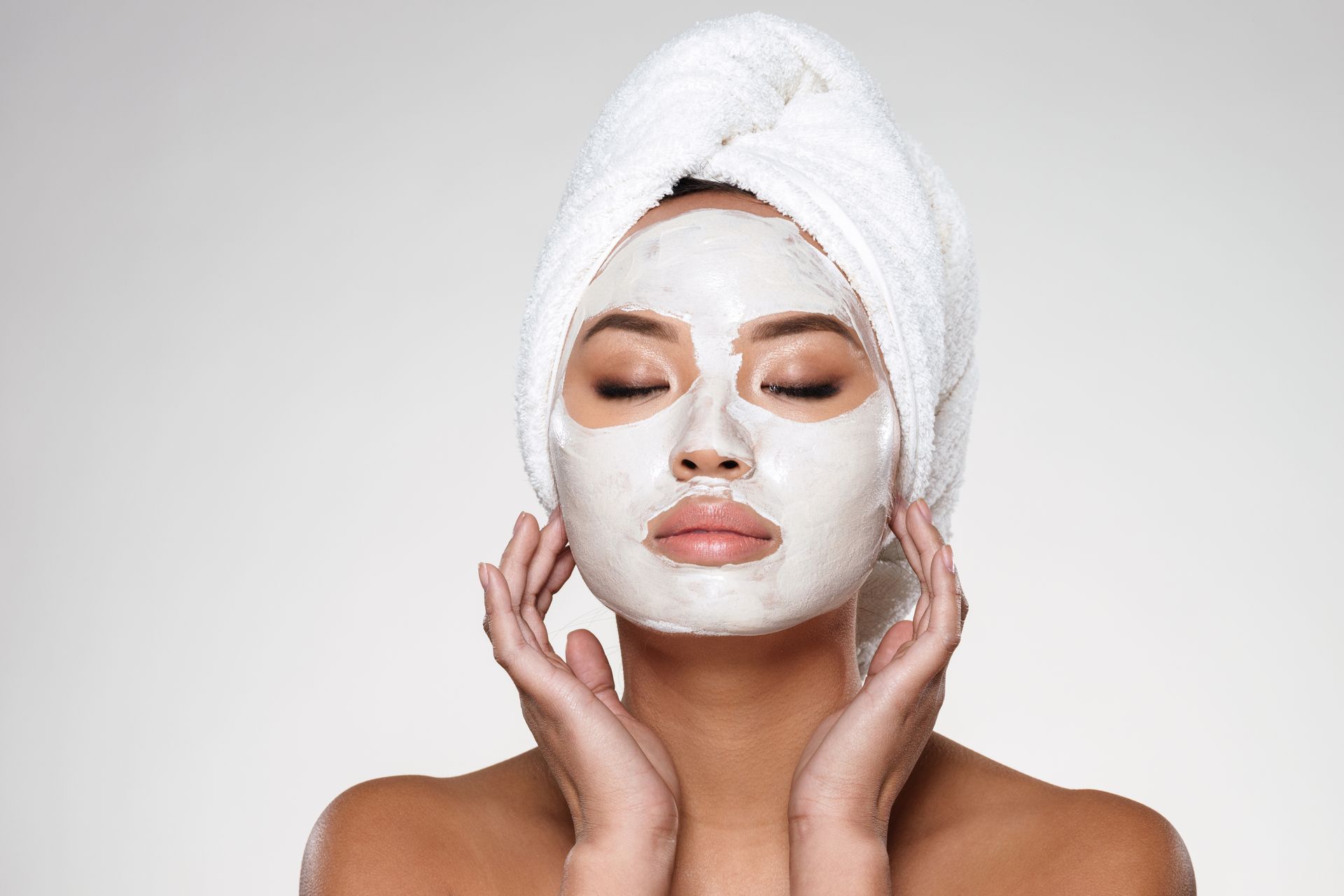 Woman with white towel and facial mask, eyes closed, gently touching her cheeks against a light background